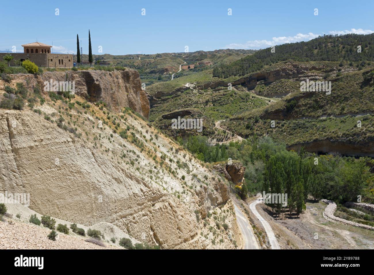 Un edificio su una scogliera rocciosa con alberi e sentieri in un paesaggio collinare, alla parete della diga dell'Embalse de Francisco Abellan, bacino idrico, fiume Fardes, L. Foto Stock