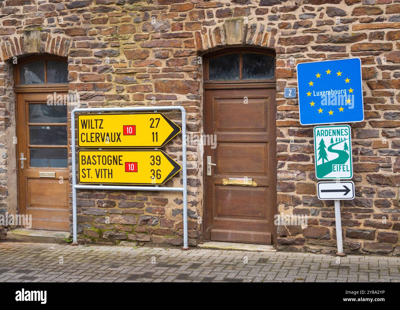 Segnale stradali gialli per Wiltz, Clervaux, Bastogne e St. Vith, con un indicatore del Lussemburgo e un cartello per sentieri, sono montati su un edificio in pietra, Lussemburgo. Foto Stock