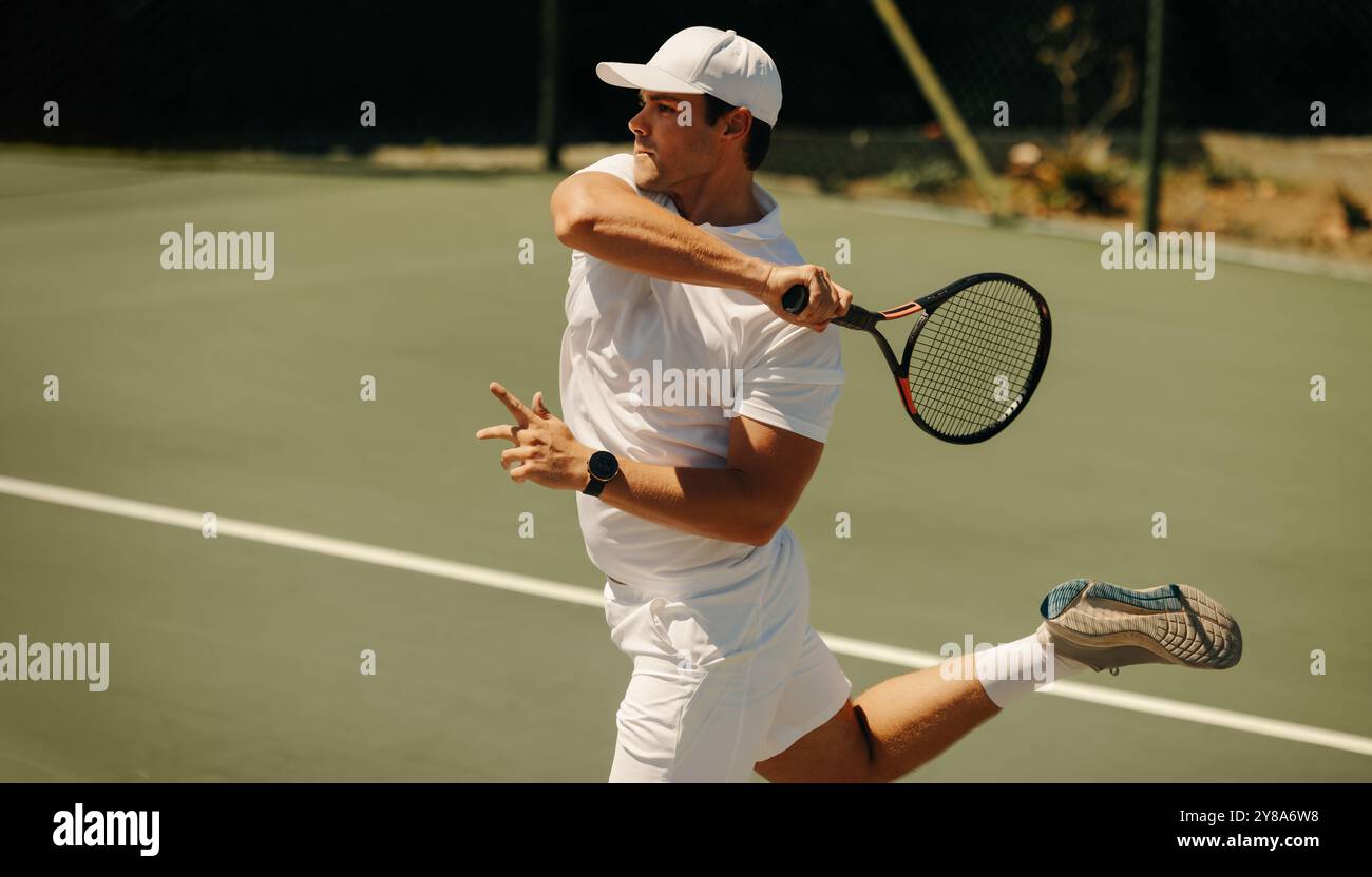 Atleta in abito bianco che dondola la sua racchetta da tennis mentre salta su un campo soleggiato all'aperto durante una partita competitiva. Foto Stock