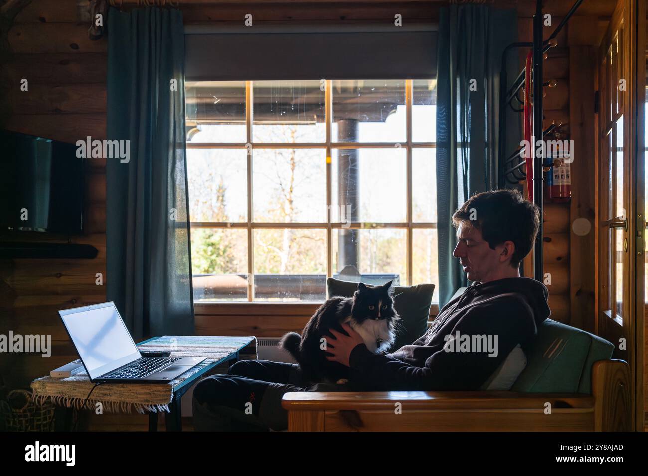 uomo seduto sul divano e con un gatto nero davanti a una grande finestra Foto Stock