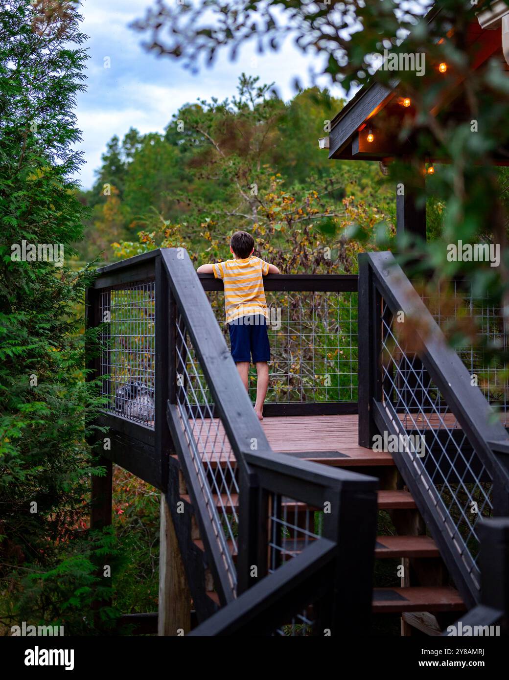 Un ragazzo in piedi sul portico anteriore di una cabina tra le montagne boscose Foto Stock