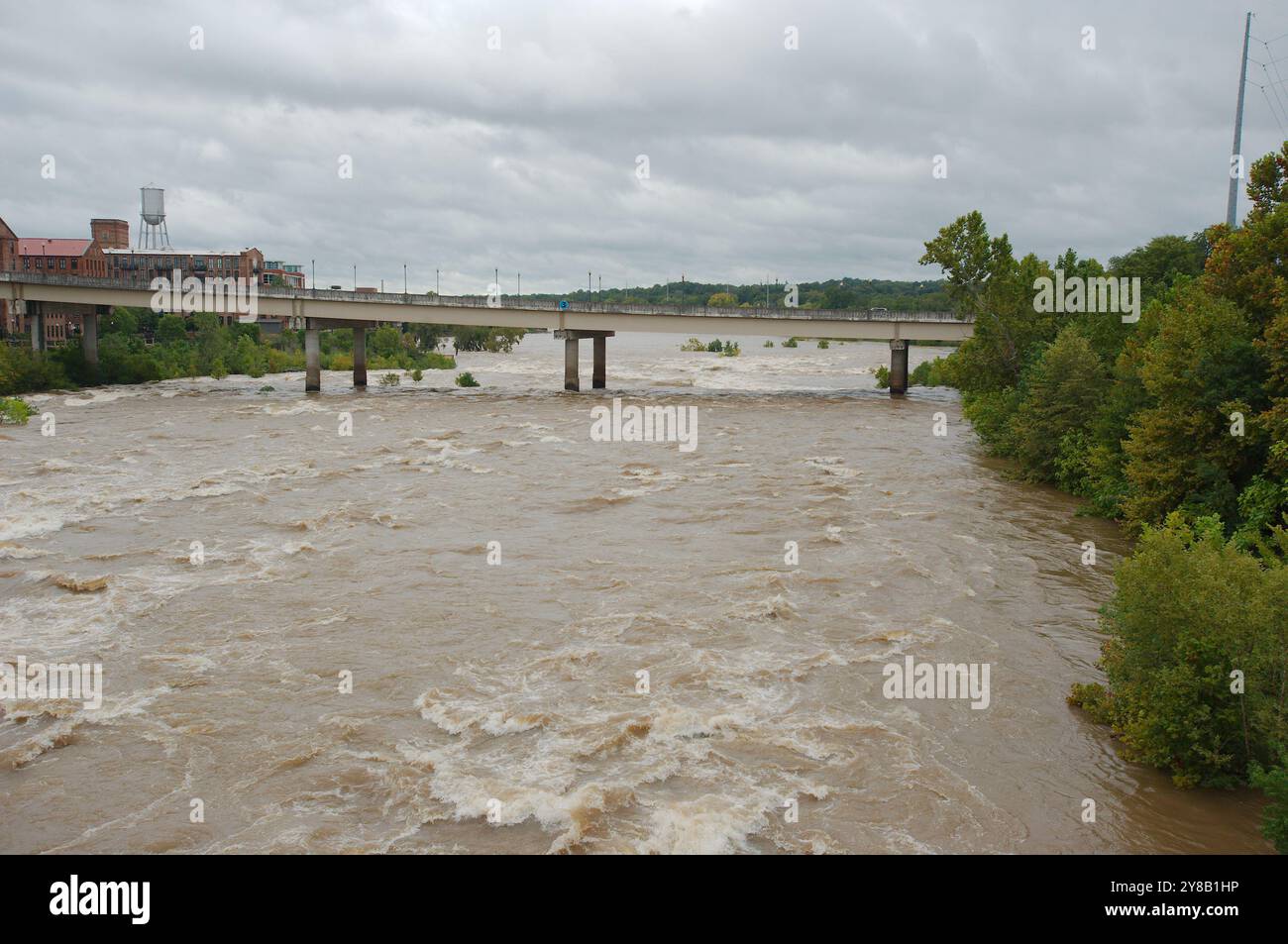 Ampia visuale dall'alto verso il basso per un rapido movimento, slancinante e fangoso flusso d'acqua di fiume marrone argilla che rotola con tappi bianchi. Alberi verdi dal ponte Foto Stock