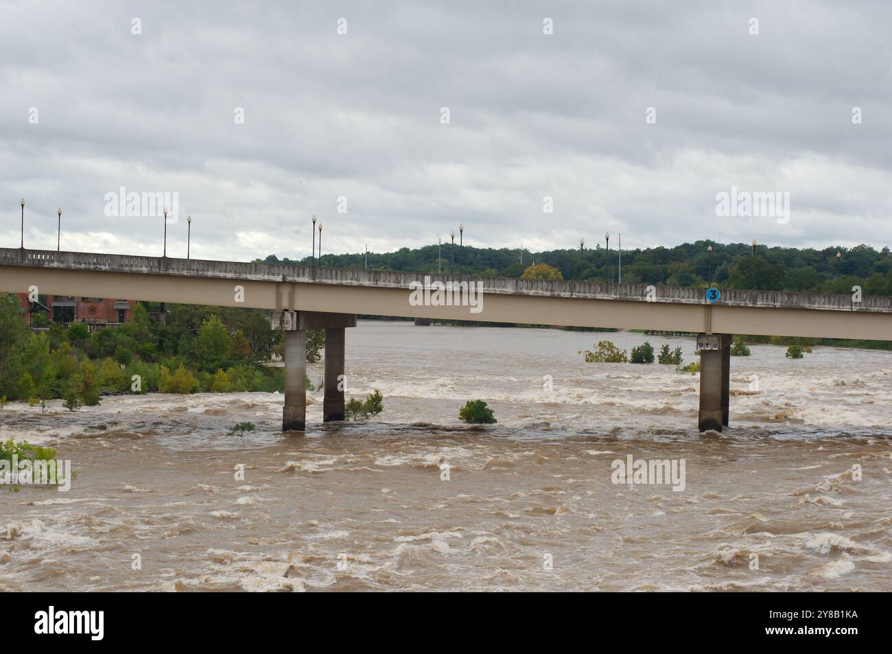 Ampia visuale dall'alto verso il basso per un rapido movimento, slancinante e fangoso flusso d'acqua di fiume marrone argilla che rotola con tappi bianchi. Alberi verdi dal ponte Foto Stock