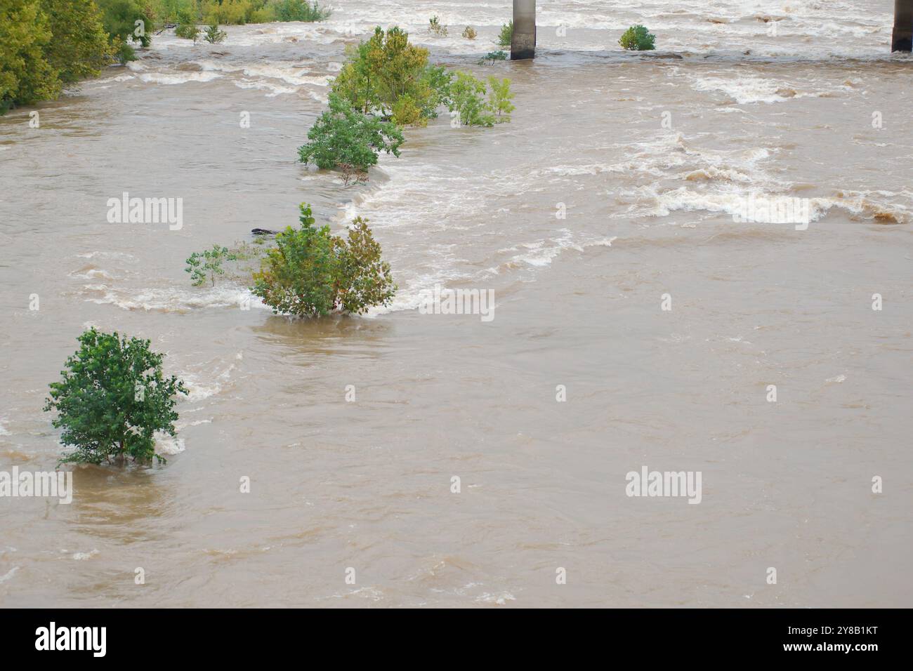 Ampia visuale dall'alto verso il basso per un rapido movimento, slancinante e fangoso flusso d'acqua di fiume marrone argilla che rotola con tappi bianchi. Alberi verdi dal ponte Foto Stock