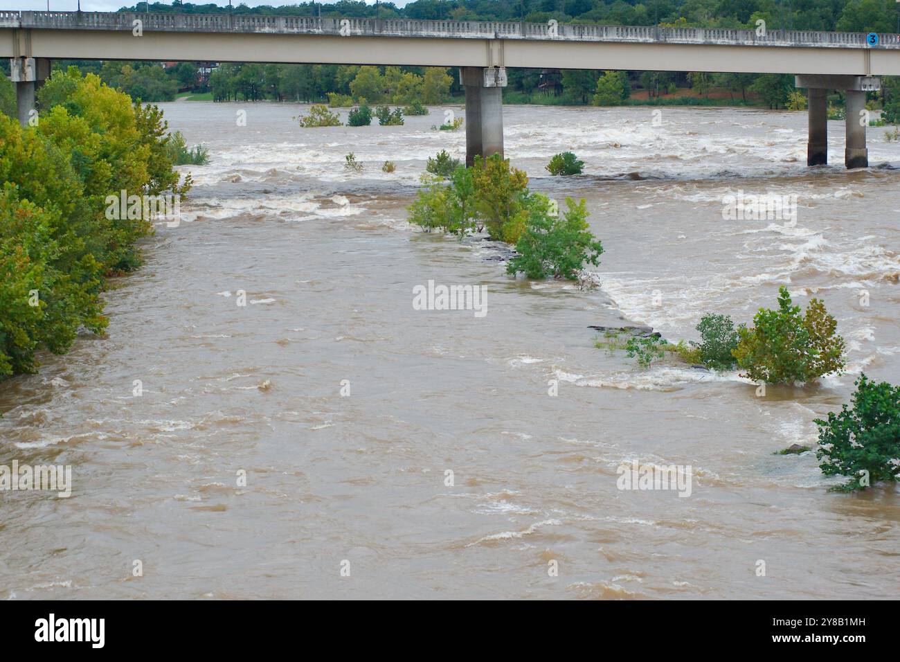 Ampia visuale dall'alto verso il basso per un rapido movimento, slancinante e fangoso flusso d'acqua di fiume marrone argilla che rotola con tappi bianchi. Alberi verdi dal ponte Foto Stock