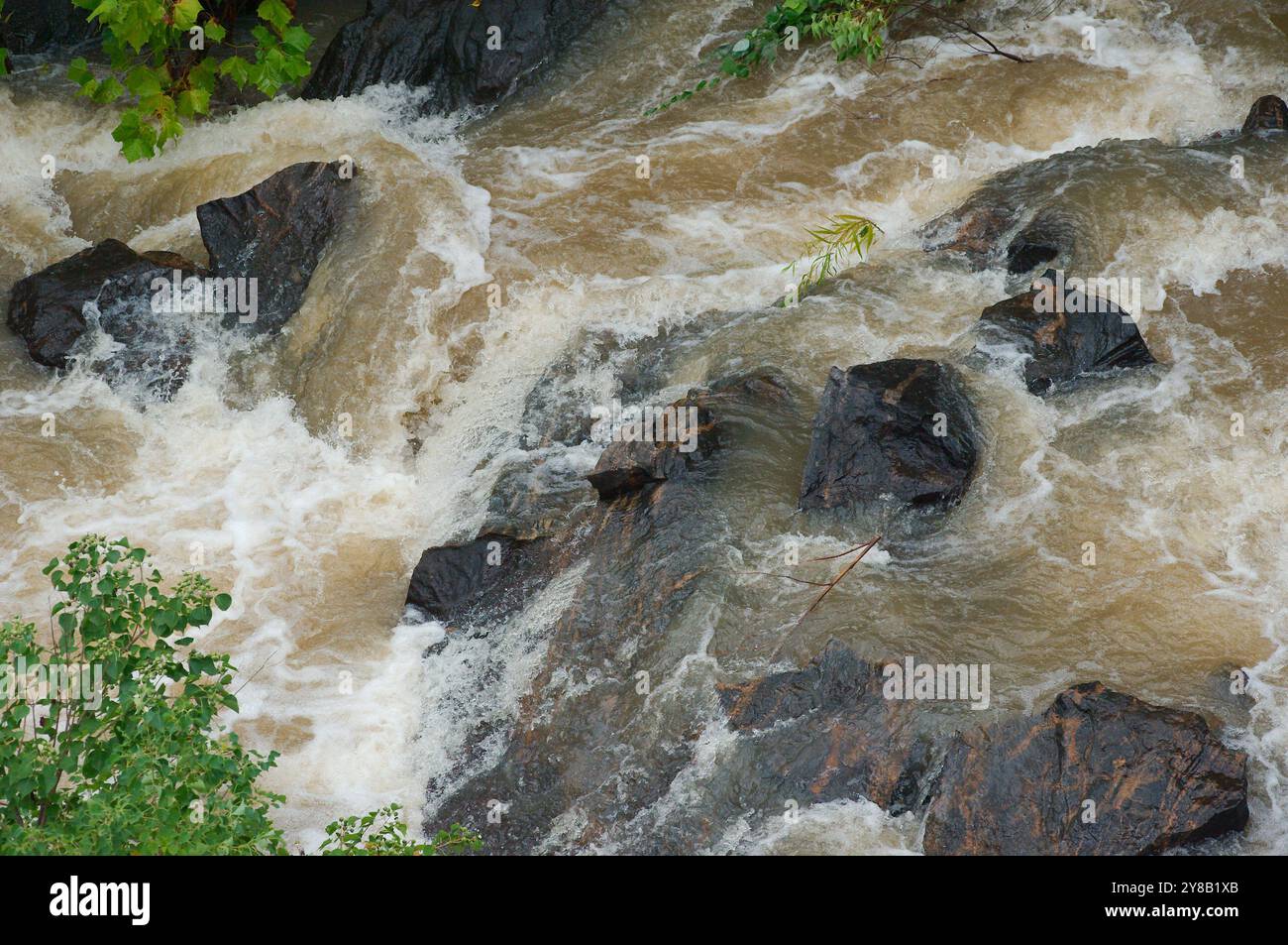 Vista ravvicinata dall'alto che guarda verso il basso le rocce del fiume in un rapido movimento, che sgorga l'acqua di un fiume marrone argilla fangosa che scorre con le berretti bianche. Aggress Foto Stock