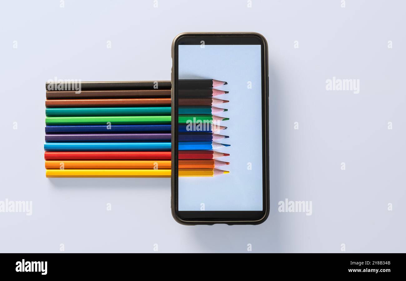 Fila di matite colorate, con punte rivolte verso destra. Poggiano su una superficie bianca, accanto a uno smartphone. Sullo schermo del telefono viene visualizzato continua Foto Stock