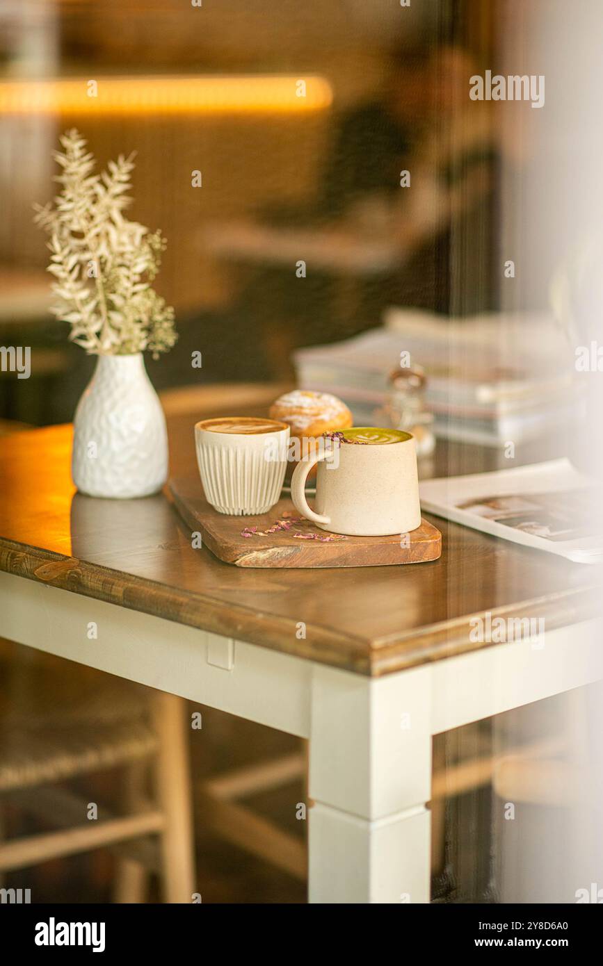 Accogliente tavolino da caffè con due tazze da caffè in ceramica, una pasticceria, un vaso bianco con fiori secchi e una rivista, inondata di calda luce naturale. Foto Stock