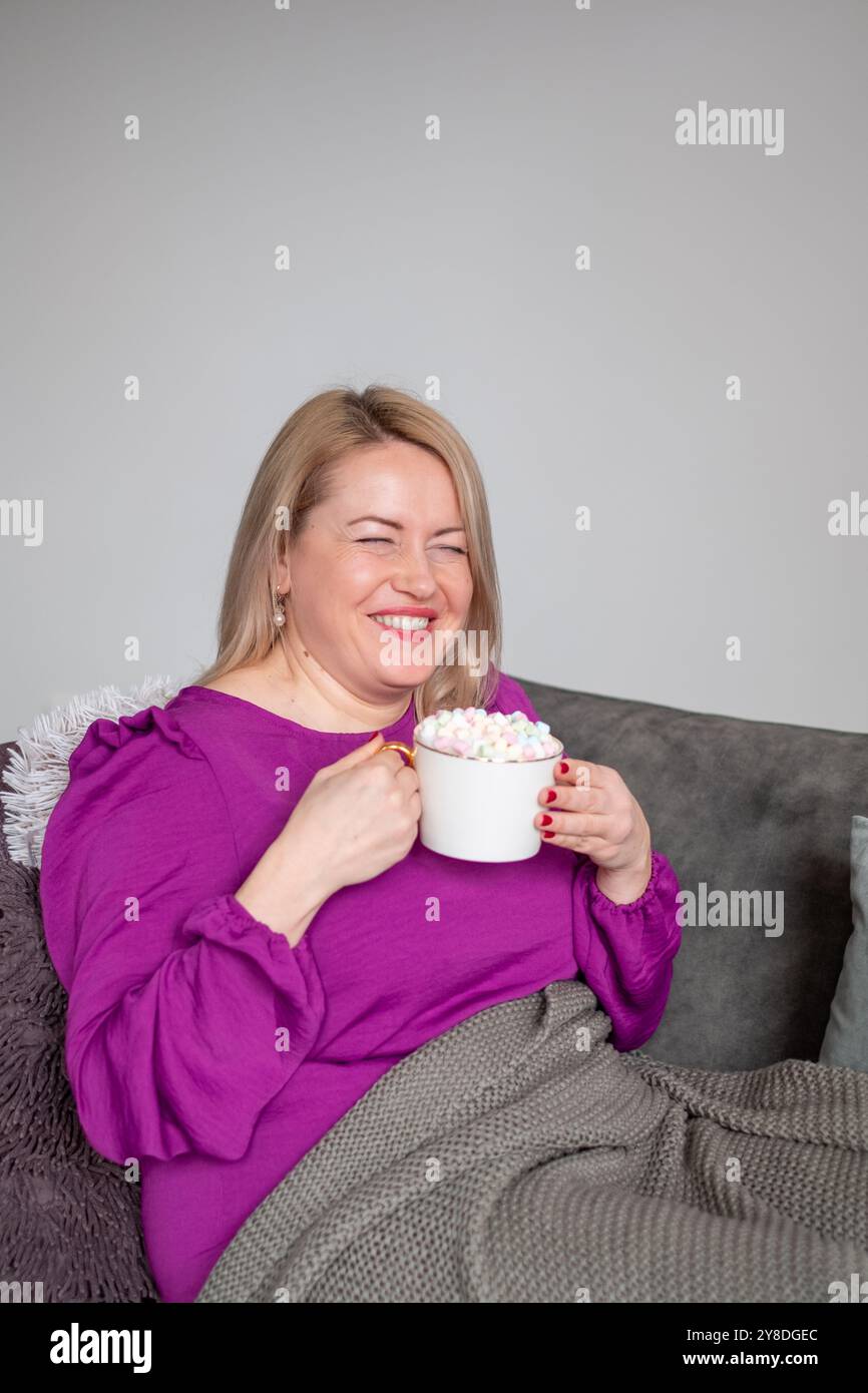 Donna rilassata che sorride sul divano tenendo in mano cioccolata calda, comoda aura in salotto, concetto di calore e intimità Foto Stock