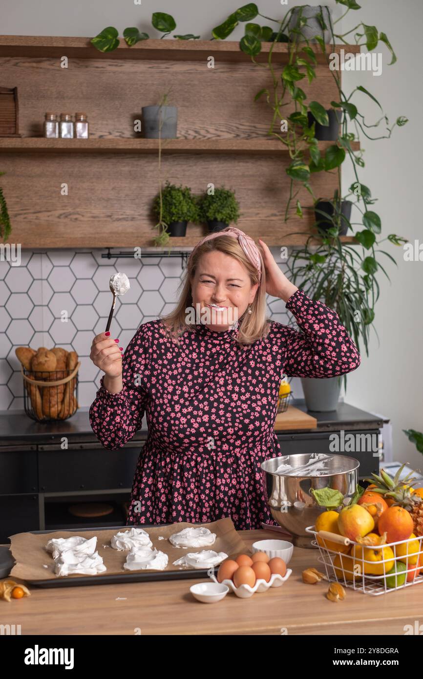 In un ambiente di cucina di casa, una donna con un abito floreale mostra un'espressione confusa mentre tiene gli albumi, cuocono gli ingredienti e cucinano Foto Stock