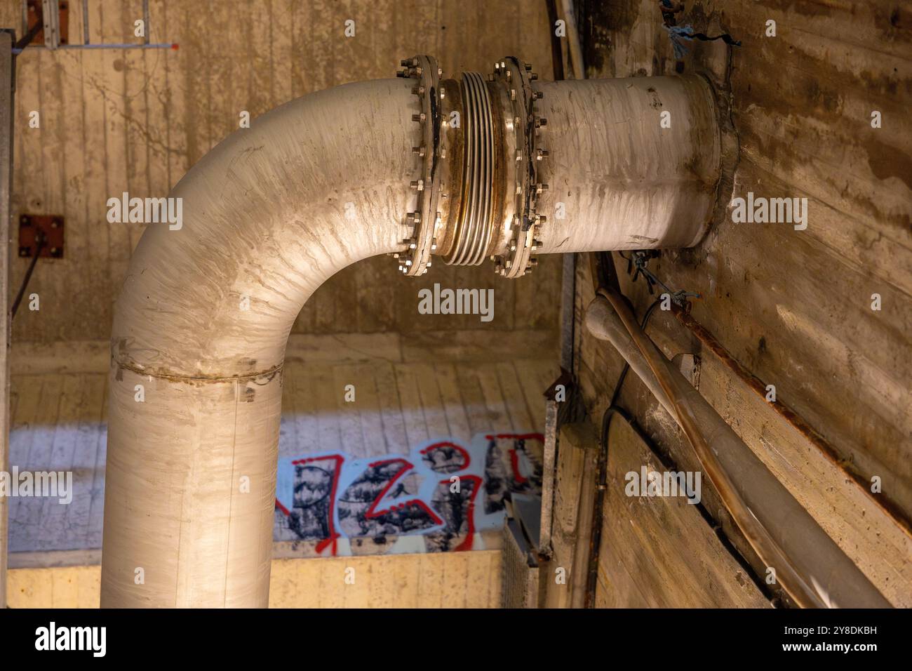 Un grande tubo industriale con un giunto metallico all'interno di una struttura in cemento, con graffiti sulle pareti. Foto Stock