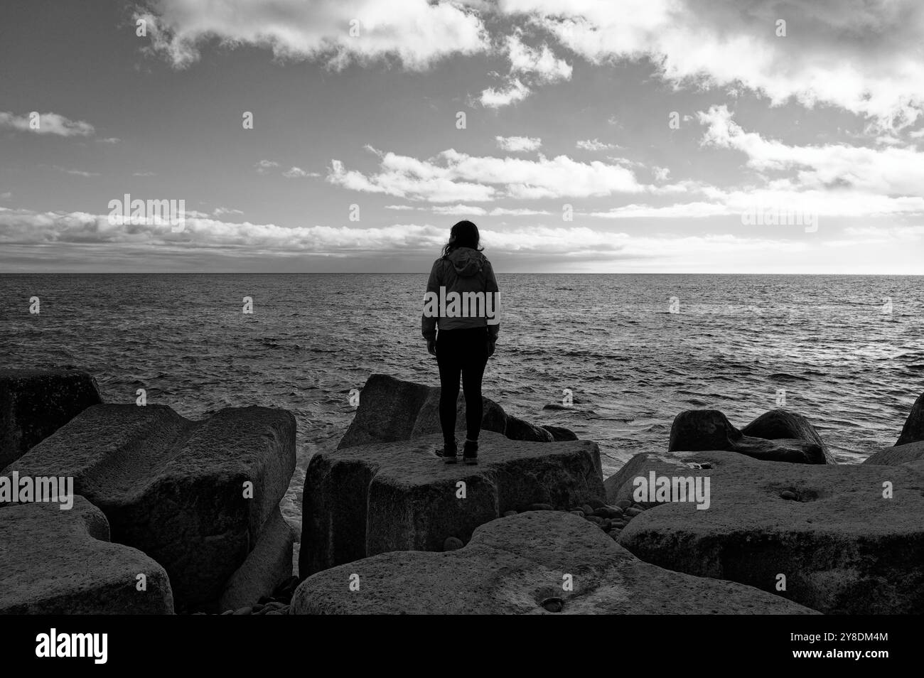 Una figura femminile solitaria guarda il vasto oceano atlantico da aspri massi artificiali costieri in bianco e nero Foto Stock