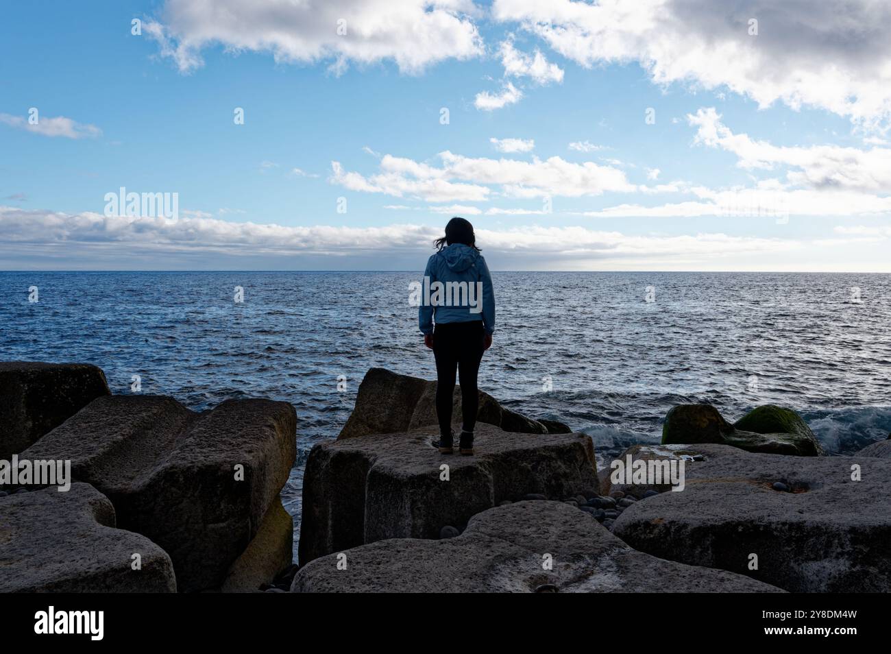 Una figura femminile solitaria si erge su grandi rocce costiere artificiali, che guardano il vasto oceano atlantico sotto un cielo blu brillante Foto Stock