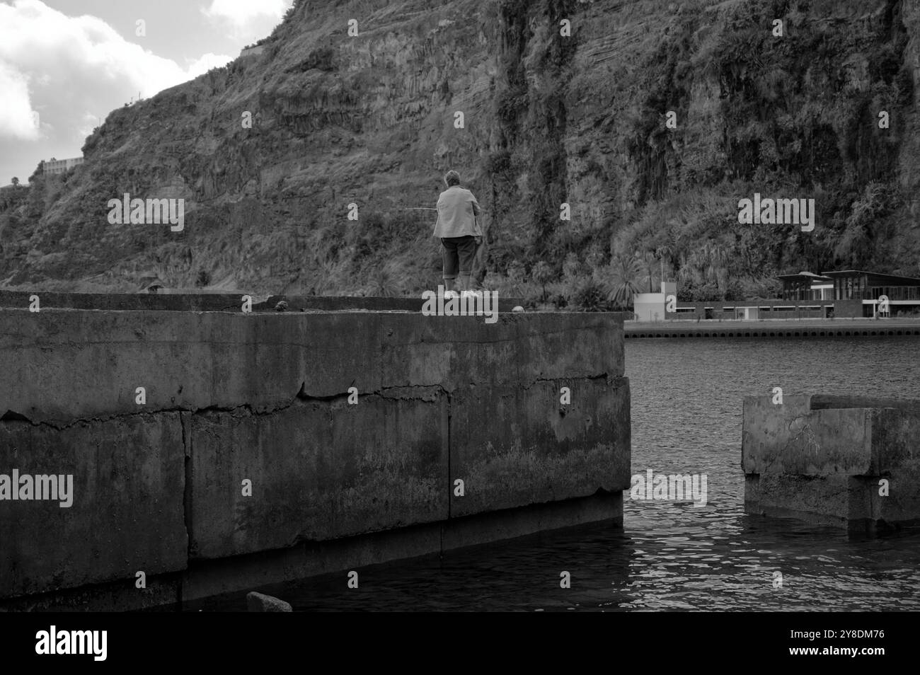 Monocromatico di una pescatrice solitaria in piedi su un aspro molo di cemento con torreggianti scogliere lungo la costa di Madeira Foto Stock