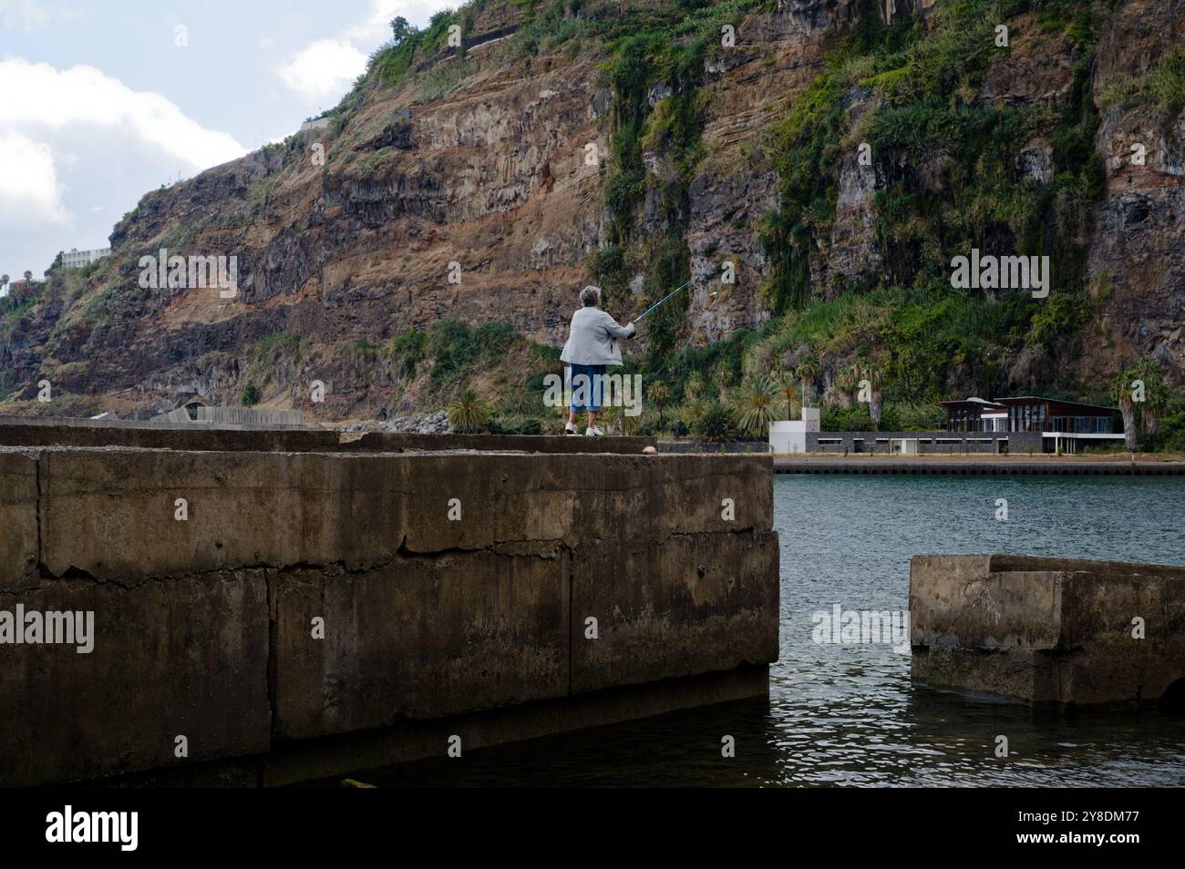 Una femmina solitaria getta la sua linea da un aspro molo di cemento, circondato da coste rocciose e scogliere lungo la spettacolare costa di Madeira Foto Stock