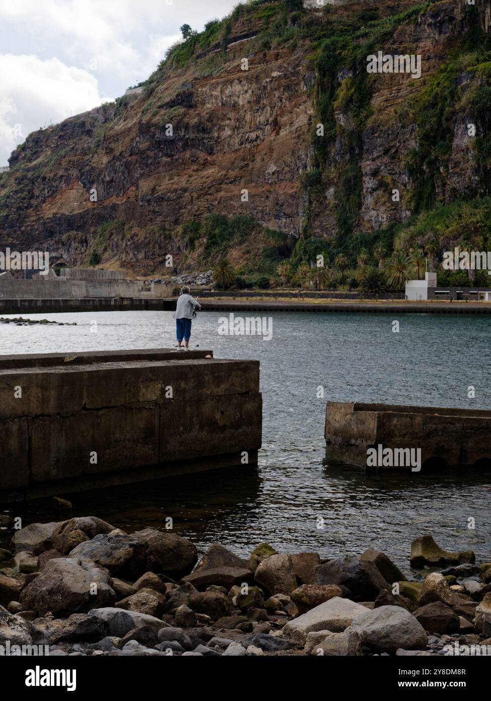 Una femmina solitaria getta la sua linea da un aspro molo di cemento, circondato da coste rocciose e scogliere lungo la spettacolare costa di Madeira Foto Stock