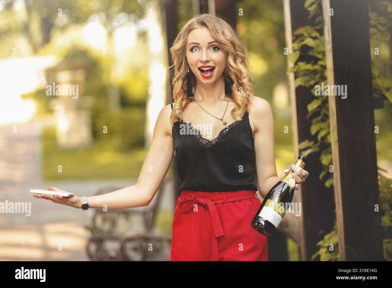 Felice giovane attraente donna bianca a ridere con gioia, parlando al telefono, tenendo in mano una bottiglia di champagne e la possibilità di chiamare un partito. emozioni lifestyle Foto Stock