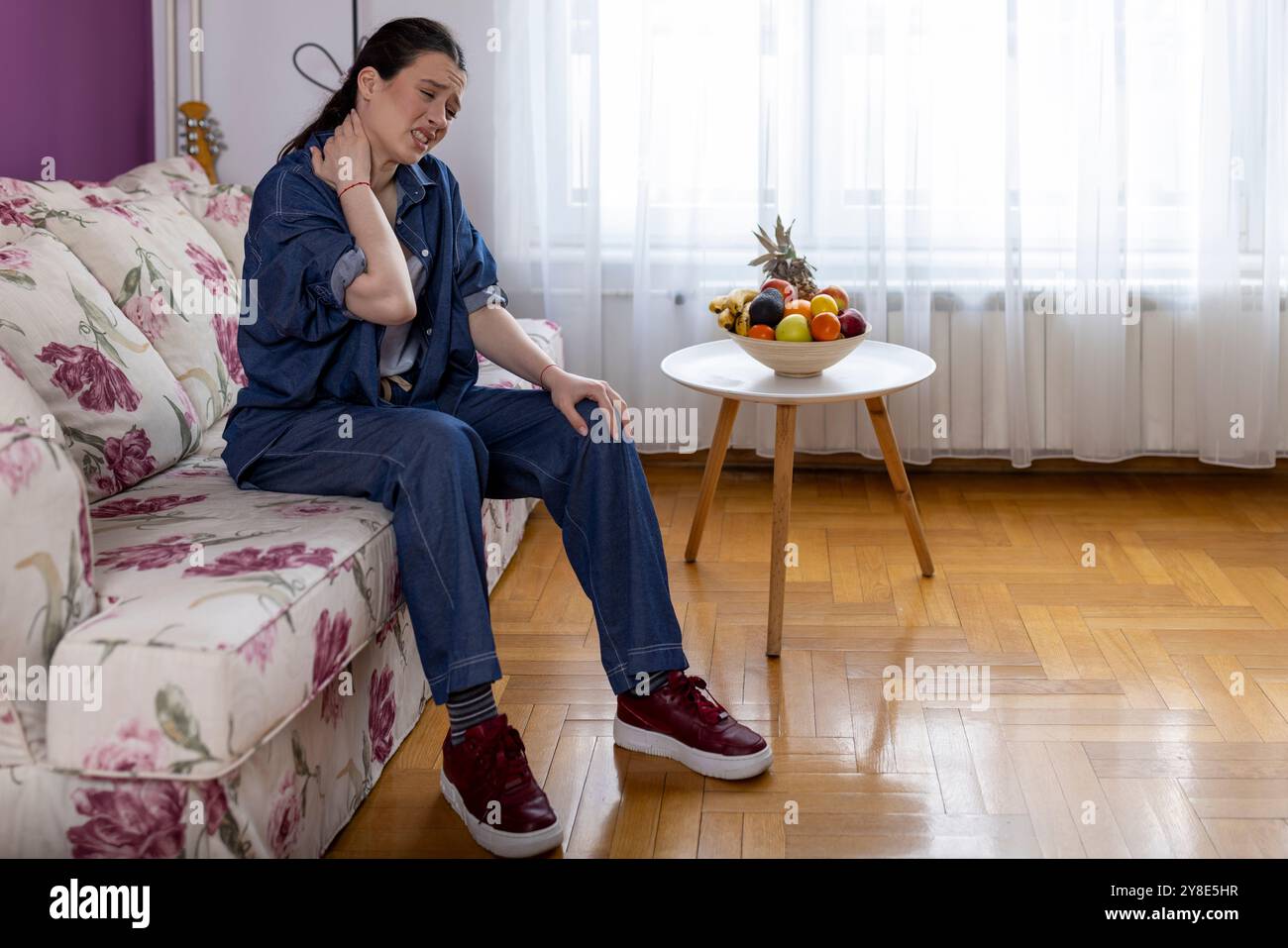 Una donna seduta sul divano con un'espressione dolorosa che tiene il collo, soffre di dolore al collo in casa, abbigliamento casual, problema di salute della donna Foto Stock