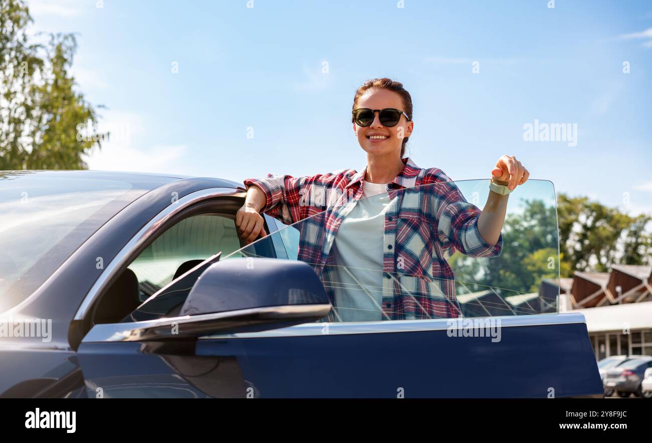 Giovane donna attraente in piedi accanto alla sua nuova auto elettrica all'aperto durante l'estate. Servizio di noleggio auto, car sharing. Foto Stock