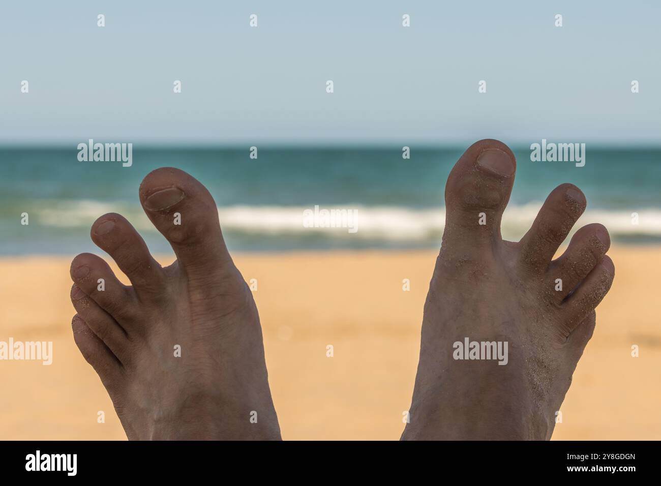 Questa immagine mostra un paio di piedi di sabbia con l'oceano e un cielo blu chiaro sullo sfondo. La scena evoca un senso di relax e divertimento, per Foto Stock