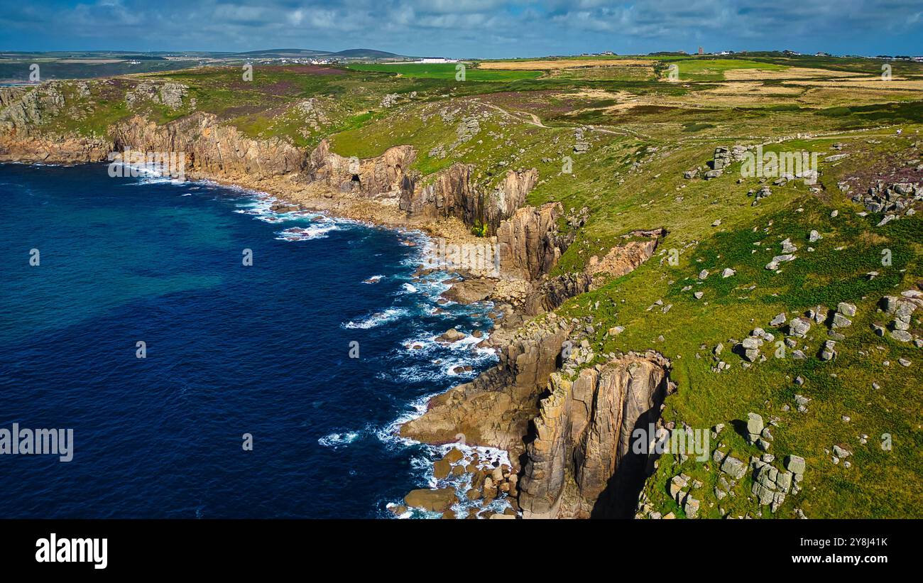 Vista aerea delle aspre scogliere costiere che incontrano il mare blu profondo, con lussureggianti campi verdi e colline ondulate sullo sfondo. La costa presenta roc Foto Stock