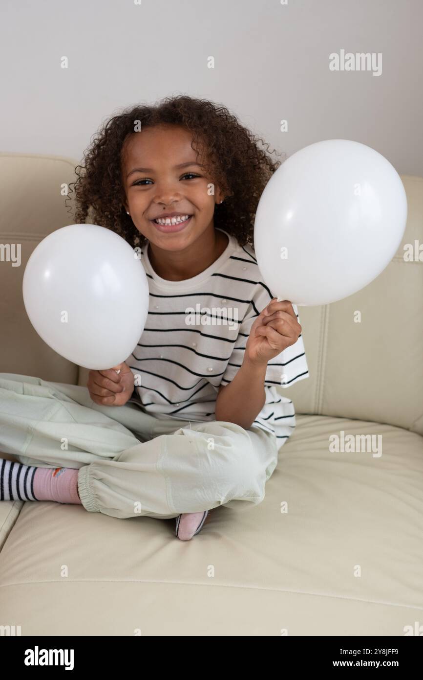 Allegro bambino seduto sul divano, con palloncini bianchi, sorridente, allegro gesto, momento giocoso, Joyous Expression, concetto di divertimento per l'infanzia Foto Stock