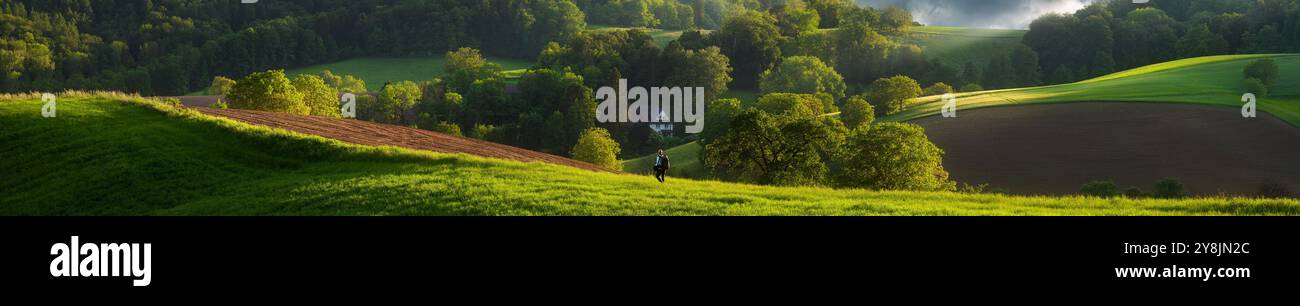 Incredibile paesaggio panoramico ultra-ampio con colline splendidamente illuminate dal sole e un escursionista sul prato verde che cammina verso una casa solitaria. Un paradiso rurale in Foto Stock