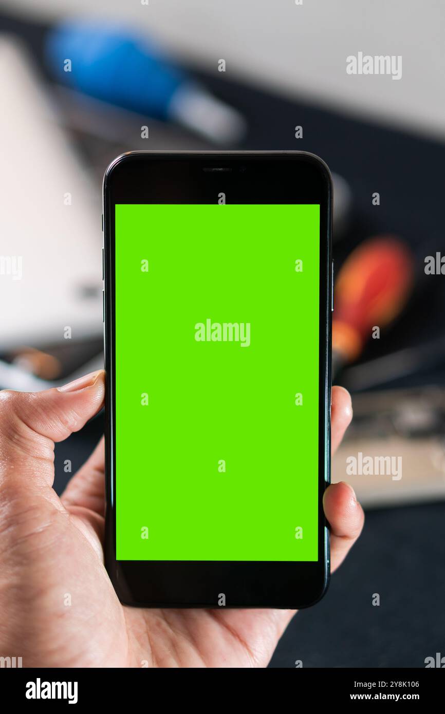 Una persona che possiede uno smartphone con schermo verde, ideale per lo sviluppo di app o la pubblicità. Lo sfondo presenta elementi da ufficio sfocati, creando un Foto Stock