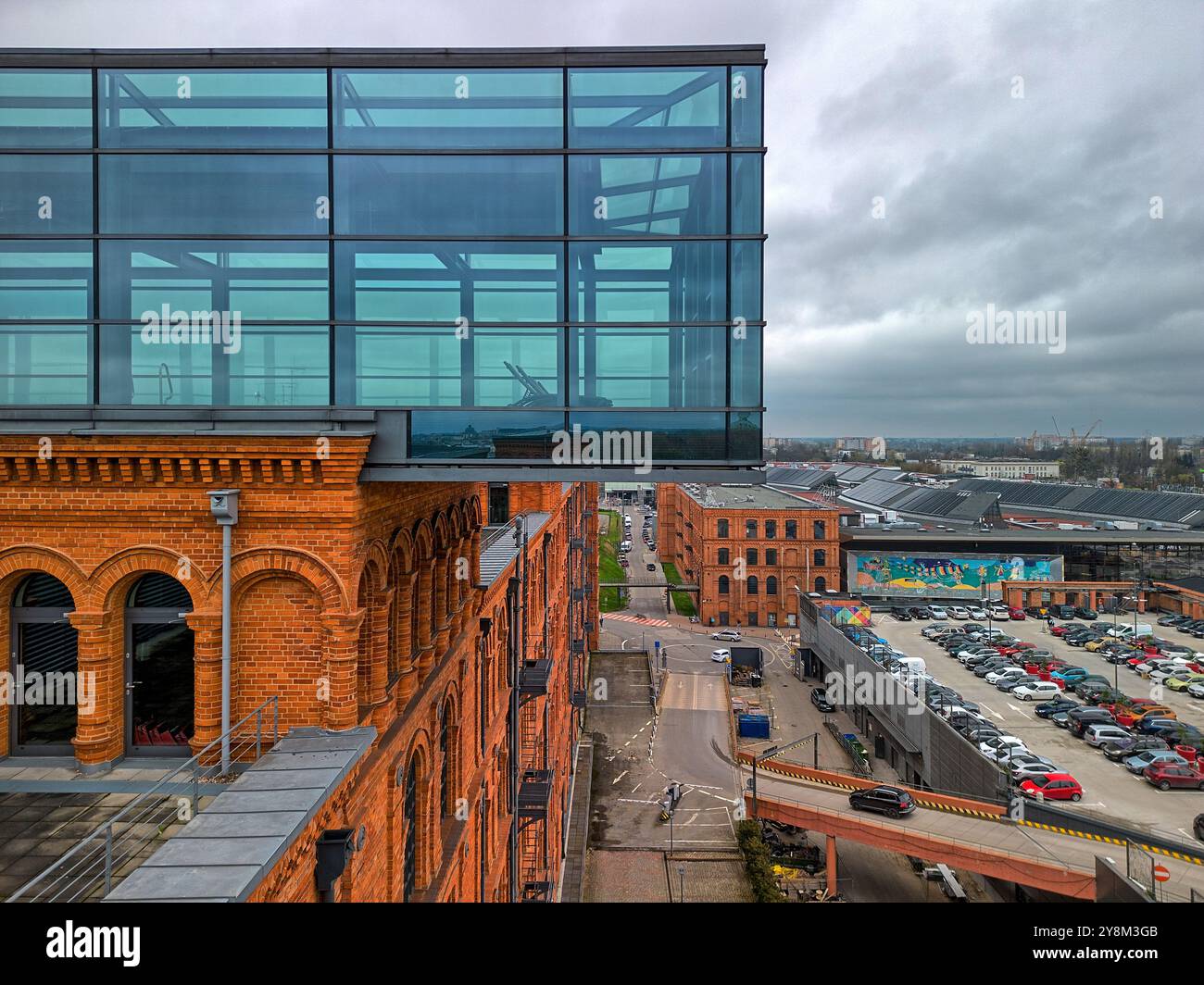Moderna struttura in vetro che si estende da un edificio industriale in mattoni rossi a Manufaktura, Łódź, Polonia, con vista sul parcheggio e sulle aree pubbliche del complesso Foto Stock