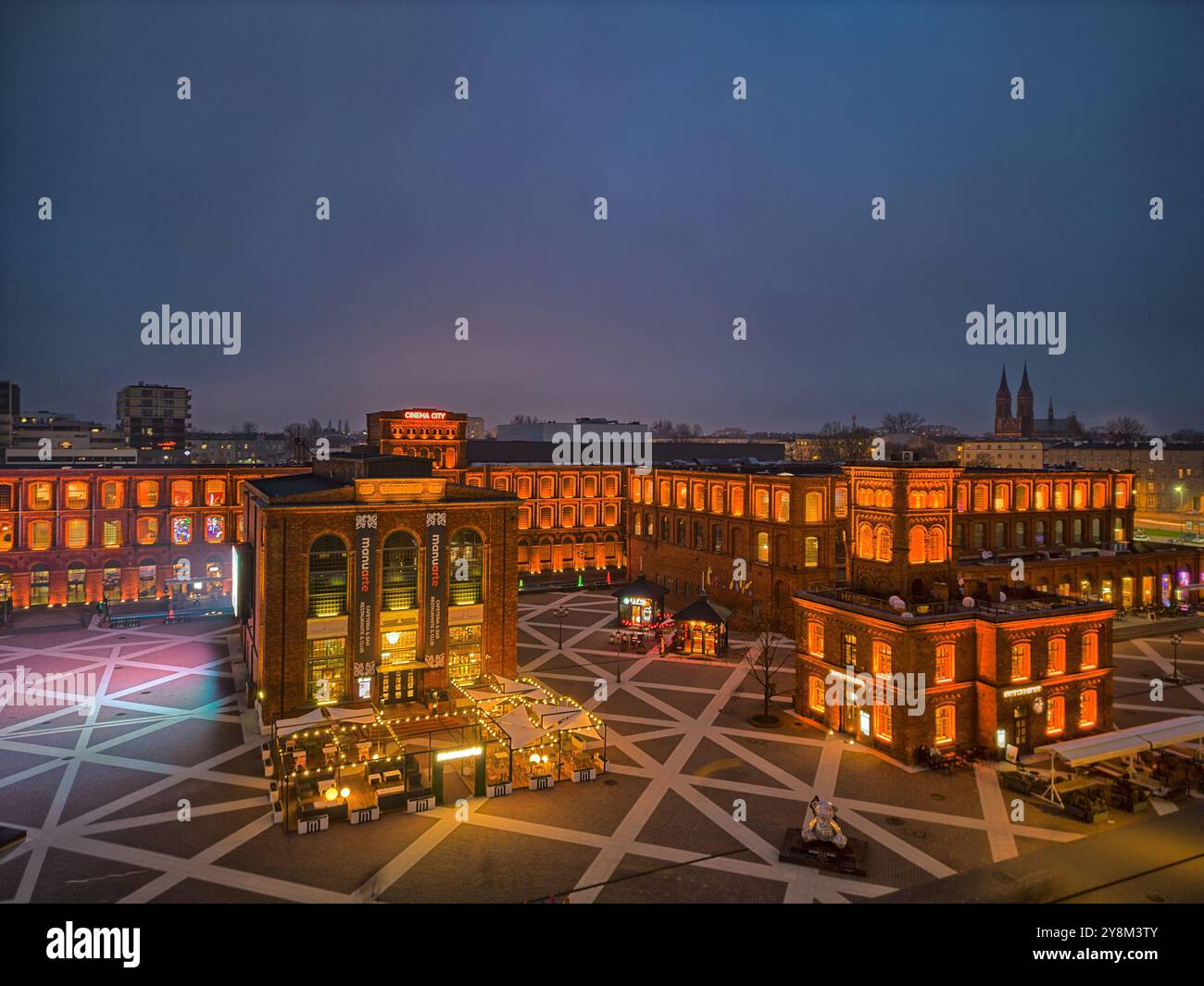 Vista notturna di Manufaktura a Łódź, Polonia, illuminata da una calda illuminazione contro gli storici edifici in mattoni rossi e la piazza pubblica aperta Foto Stock