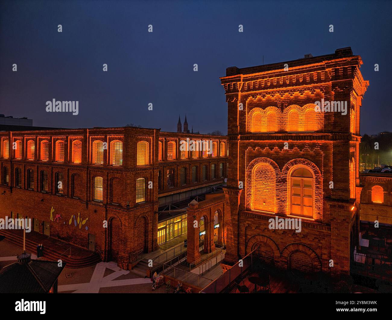 Vista notturna di Manufaktura a Łódź, Polonia, illuminata da una calda illuminazione contro gli storici edifici in mattoni rossi e la piazza pubblica aperta Foto Stock