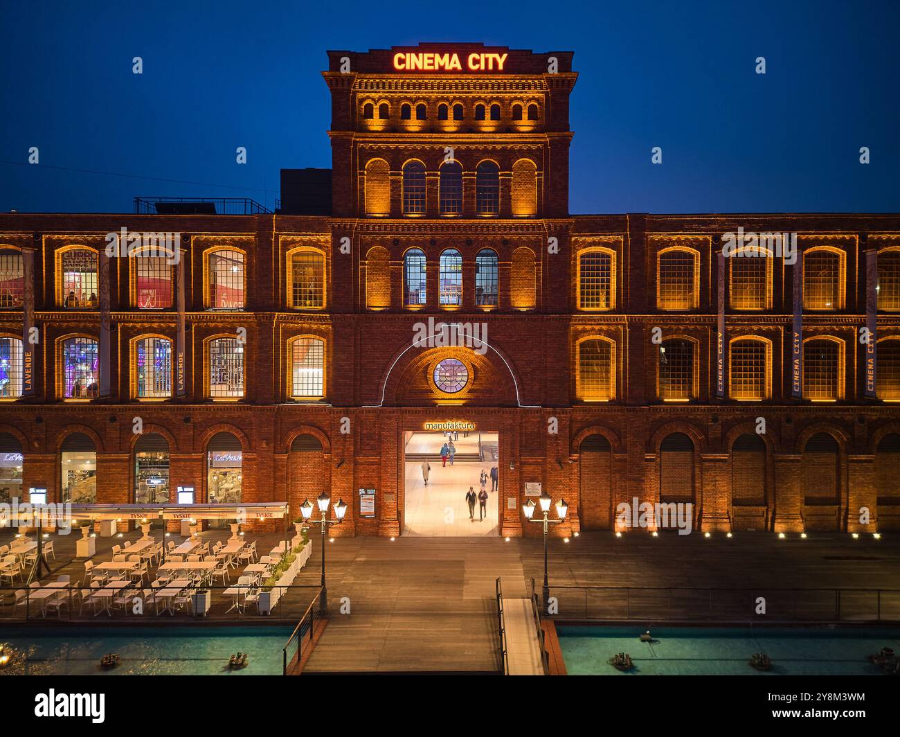 Vista notturna di Manufaktura a Łódź, Polonia, illuminata da una calda illuminazione contro gli storici edifici in mattoni rossi e la piazza pubblica aperta Foto Stock