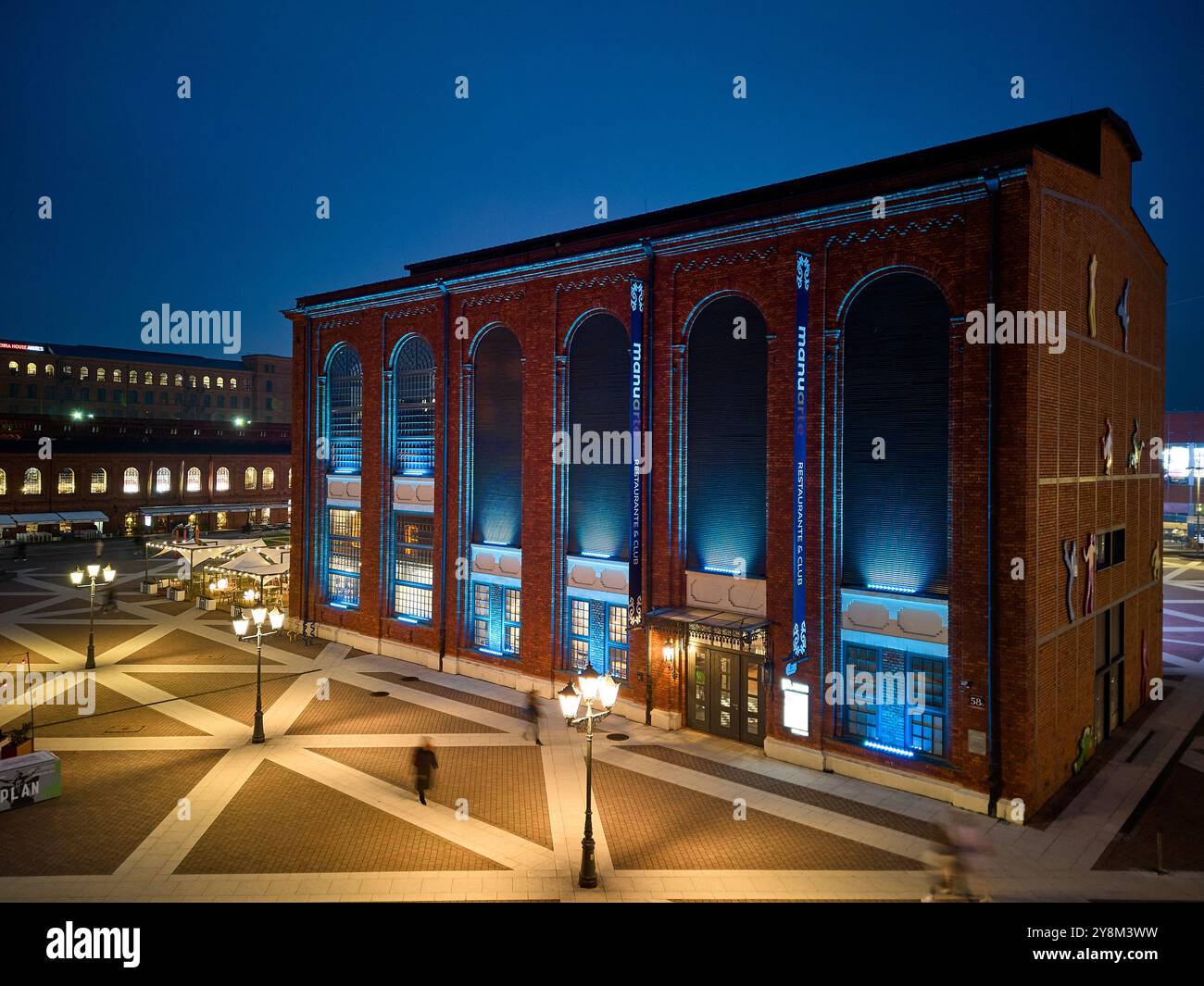 Vista notturna di Manufaktura a Łódź, Polonia, illuminata da una calda illuminazione contro gli storici edifici in mattoni rossi e la piazza pubblica aperta Foto Stock