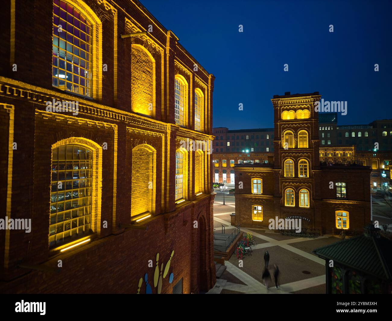 Vista notturna di Manufaktura a Łódź, Polonia, illuminata da una calda illuminazione contro gli storici edifici in mattoni rossi e la piazza pubblica aperta Foto Stock