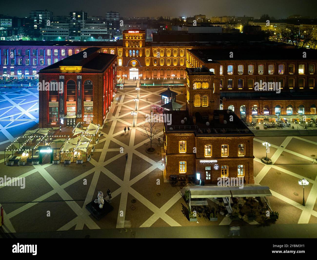 Vista notturna di Manufaktura a Łódź, Polonia, illuminata da una calda illuminazione contro gli storici edifici in mattoni rossi e la piazza pubblica aperta Foto Stock