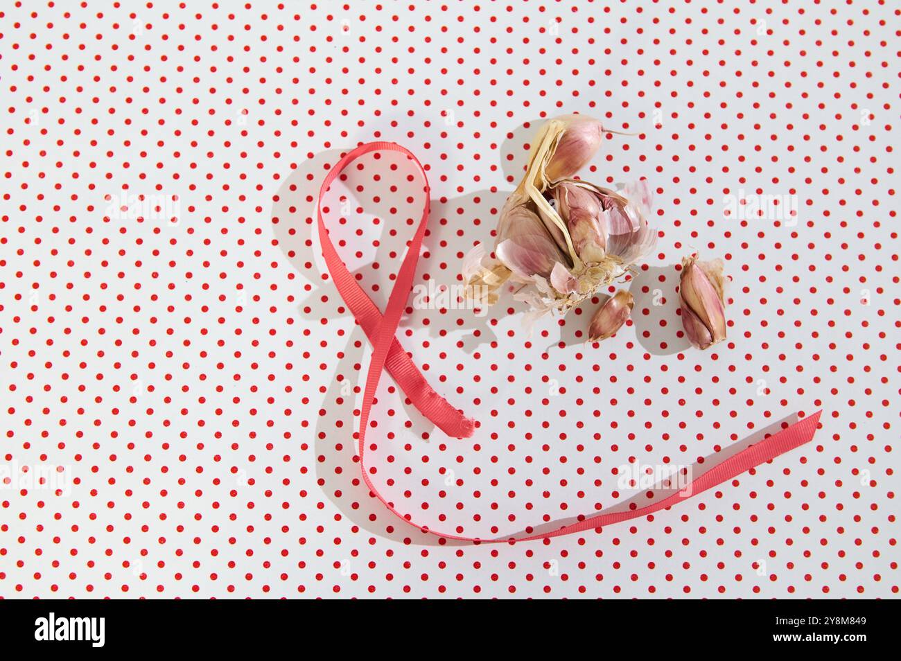 Nastro rosa accanto a bulbi d'aglio su uno sfondo a pois rosso, simboleggia la semplicità culinaria con fascino visivo. Foto Stock