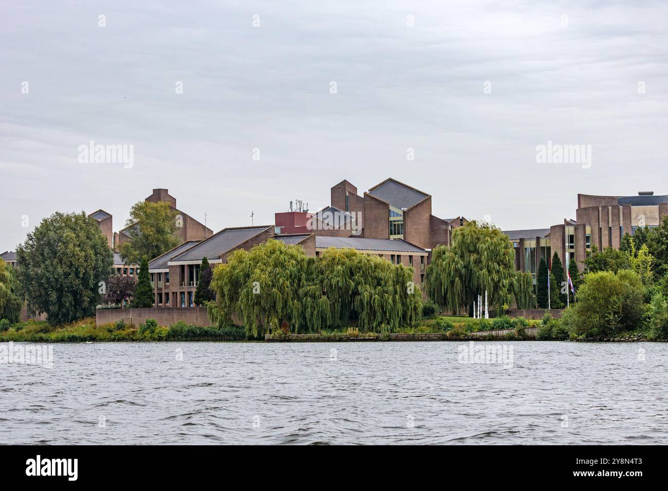 Gouvernement aan de Maas contro il cielo grigio, edificio governativo sul fiume Mosa circondato da alberi frondosi, visto da una barca, giornata nuvolosa a Maastricht, S. Foto Stock