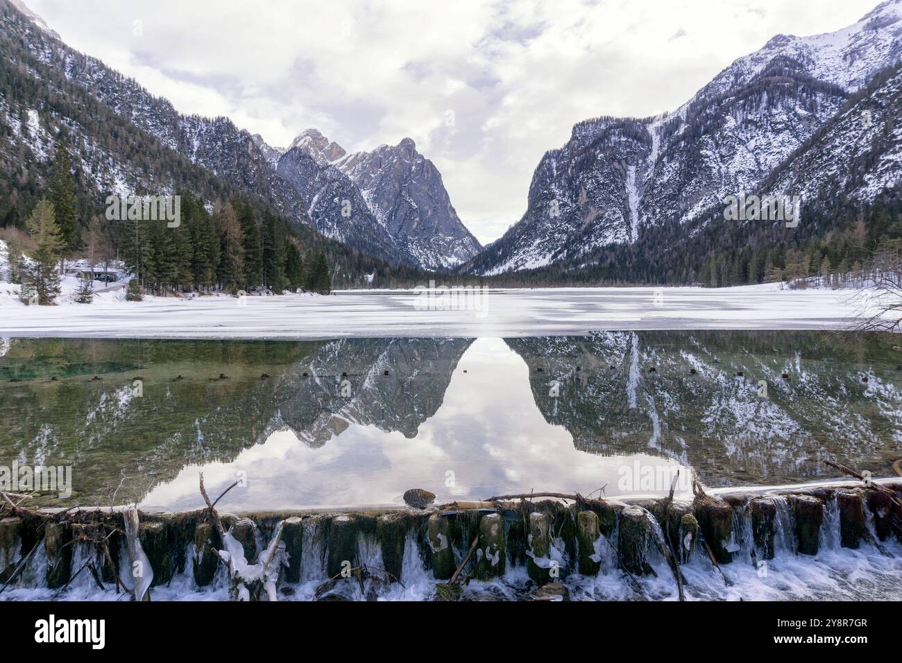 Paesaggio montano invernale in un lago di Dobbiaco innevato e parzialmente ghiacciato, montagne che si riflettono nell'acqua, cielo nuvoloso. Provincia di Bolzano, Trentino alto Foto Stock