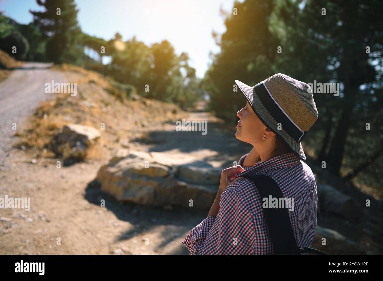 Una donna che indossa un cappello gode di una passeggiata tranquilla e soleggiata su un sentiero naturalistico panoramico, circondato da alberi e tranquillità. Foto Stock