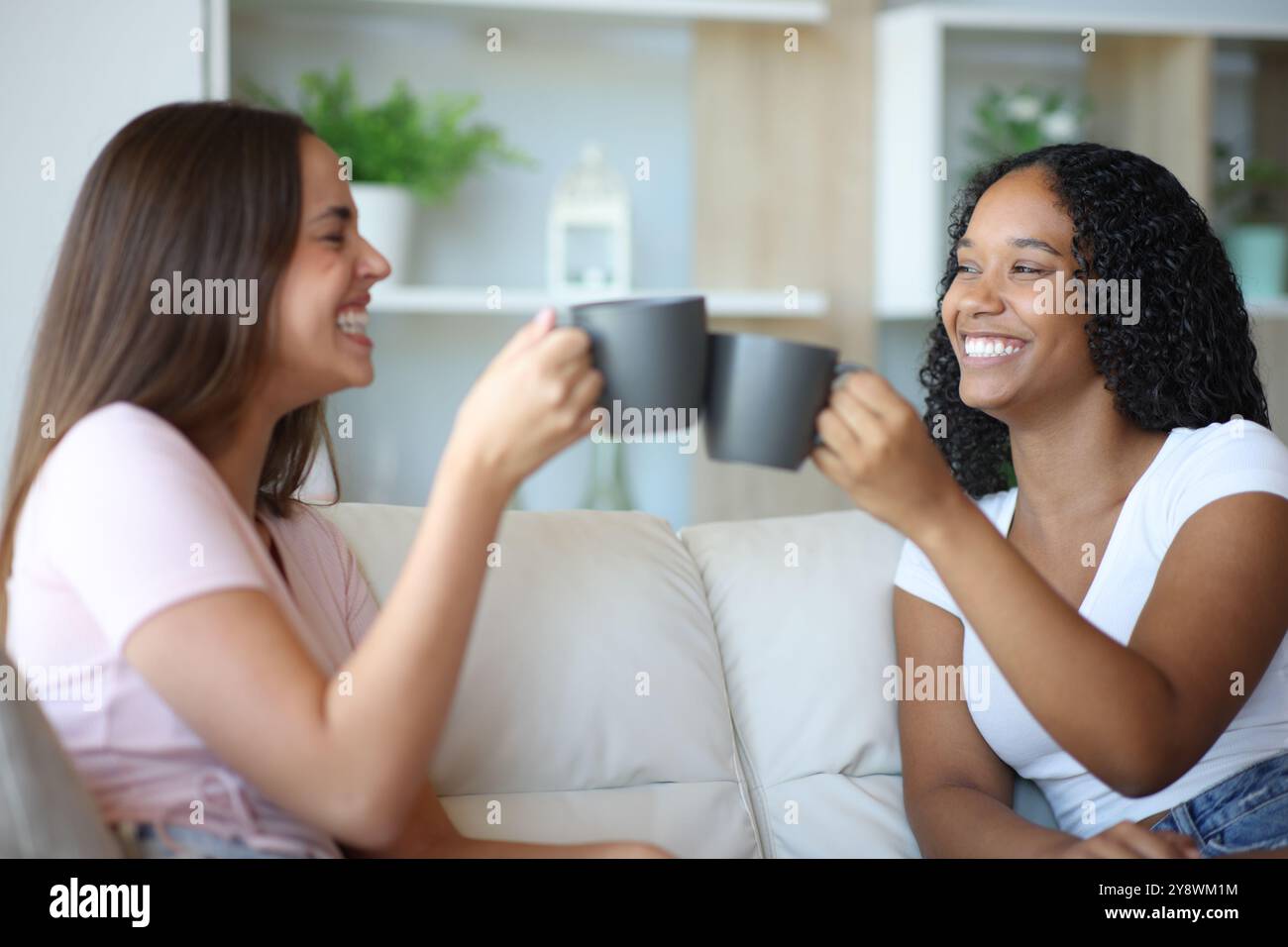 Felici compagni di stanza interrazziali che brindano parlando seduti su un divano a casa Foto Stock