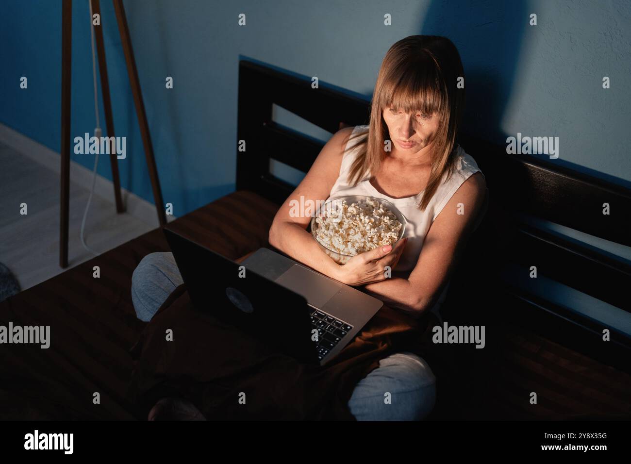 La bella donna ama i popcorn mentre guarda un film sul suo computer portatile, seduto comodamente sul suo letto di notte nella camera da letto di casa. La scena si riflette Foto Stock