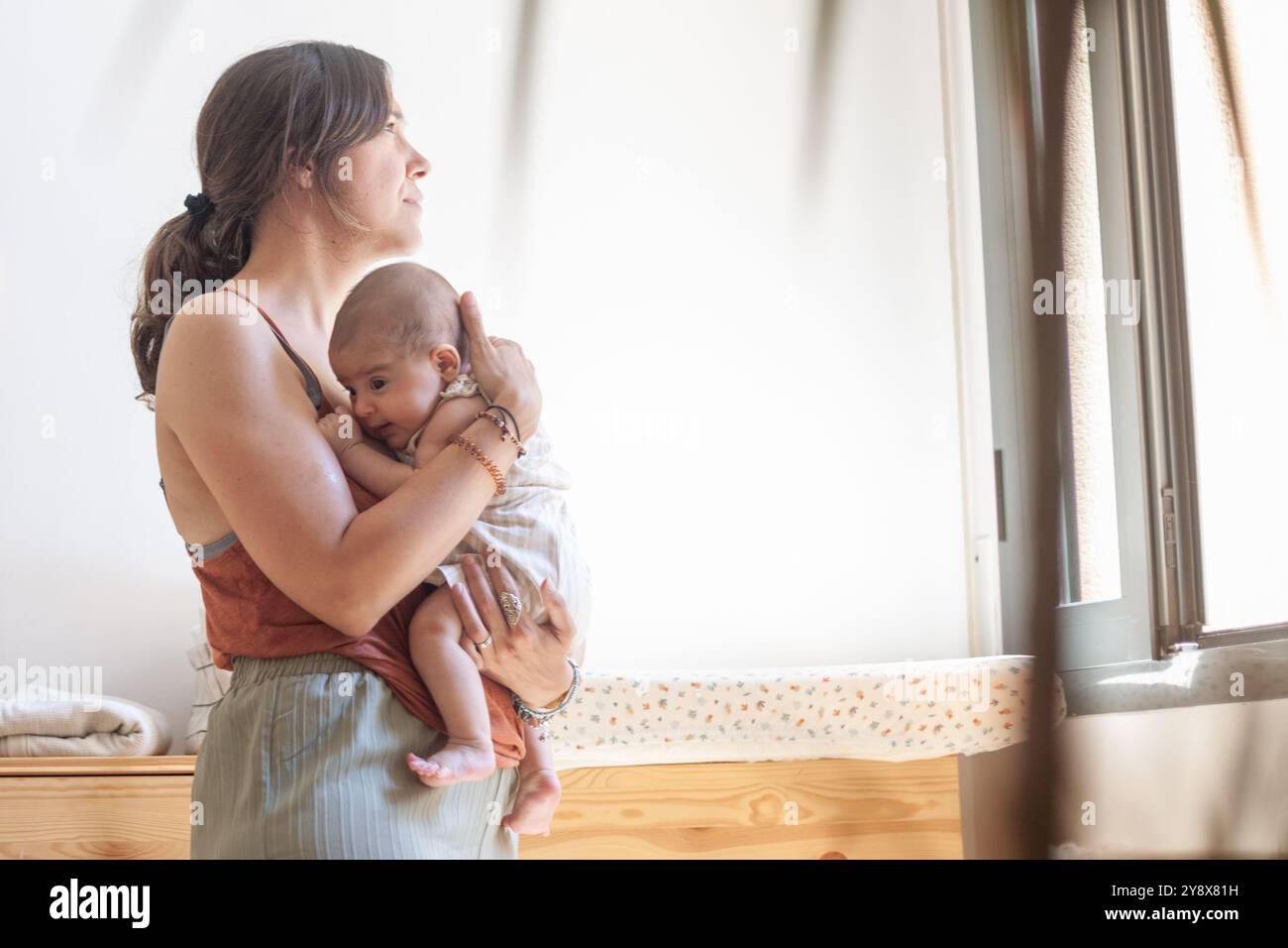 Una donna tiene un bambino tra le braccia davanti a una finestra. La scena è calda e amorevole, con la madre che fornisce conforto e cura al suo bambino. Foto Stock