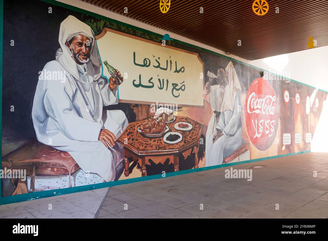 Un murale di Street art raffigura un tradizionale abito marocchino che beve bibite gassate, con testo in arabo sopra e loghi commerciali sulla destra Foto Stock