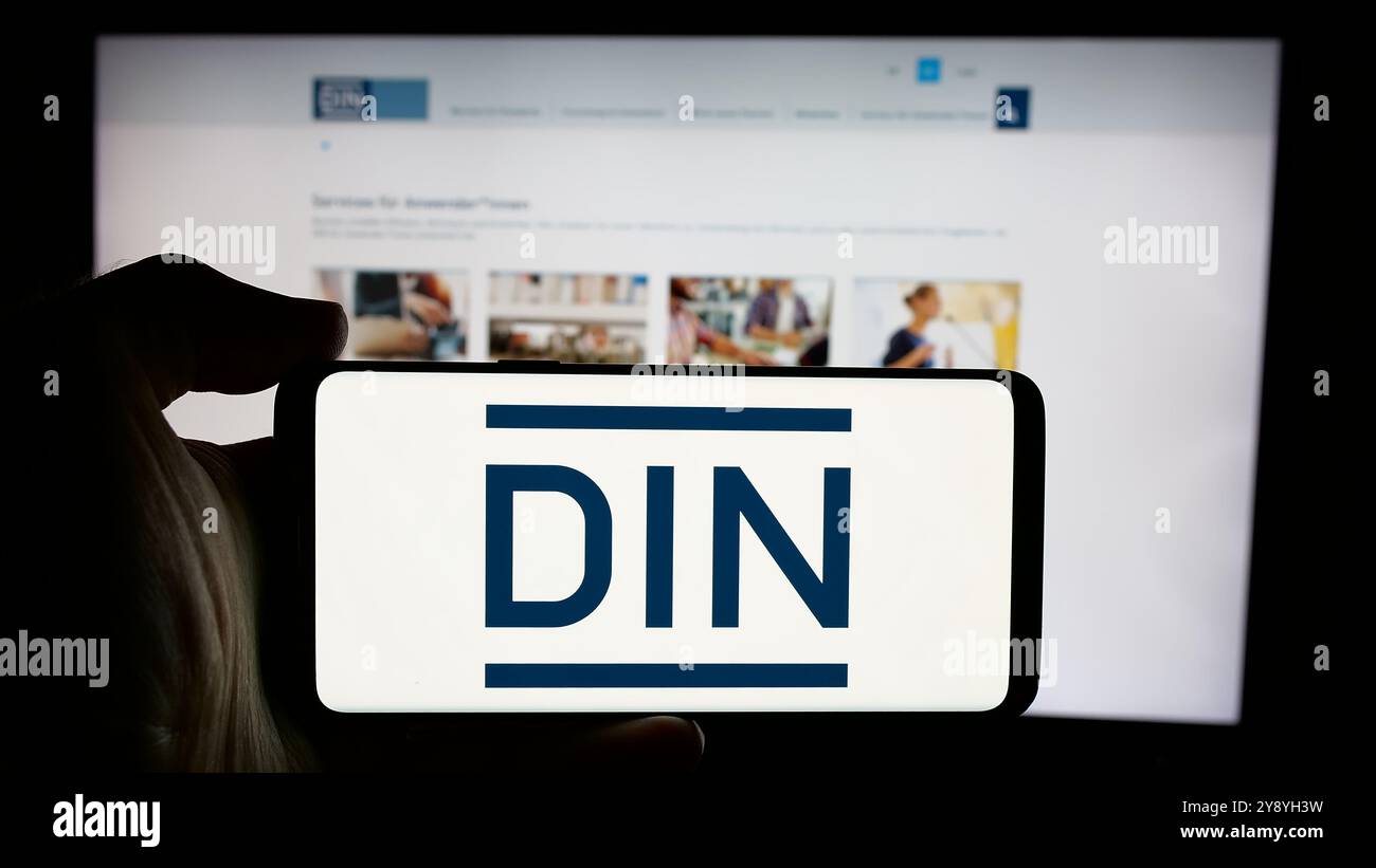 Persona che detiene un cellulare con il logo del Deutsches Institut für Normung e.V. (DIN) sullo schermo davanti alla pagina web. Mettere a fuoco il display del telefono. Foto Stock