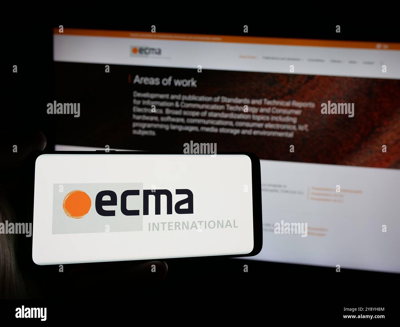 Persona che possiede uno smartphone con il logo dell'organizzazione per gli standard Ecma International sullo schermo di fronte al sito Web. Mettere a fuoco il display del telefono. Foto Stock
