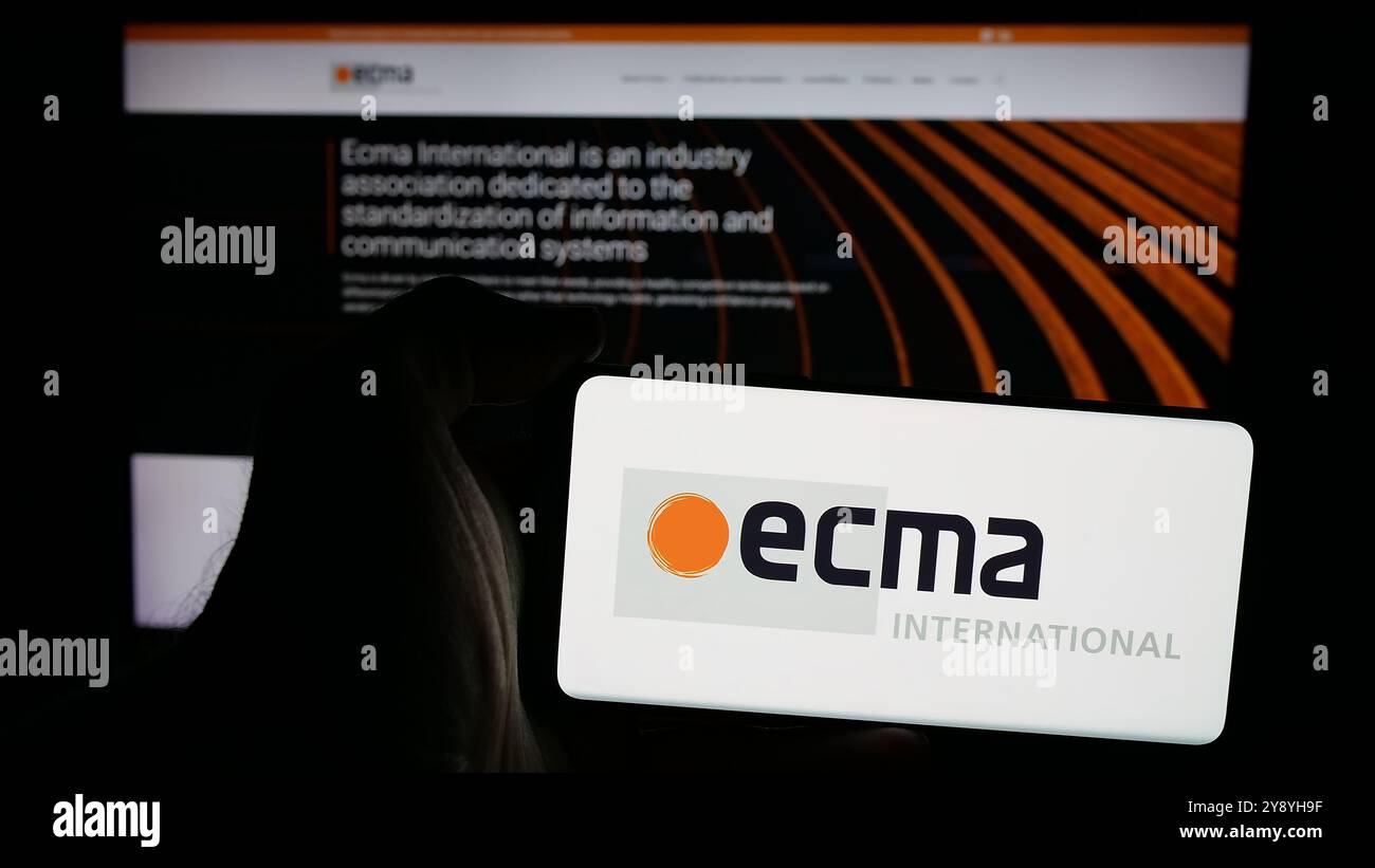 Persona che possiede un telefono cellulare con il logo dell'organizzazione di standardizzazione Ecma International sullo schermo di fronte alla pagina Web. Mettere a fuoco il display del telefono. Foto Stock