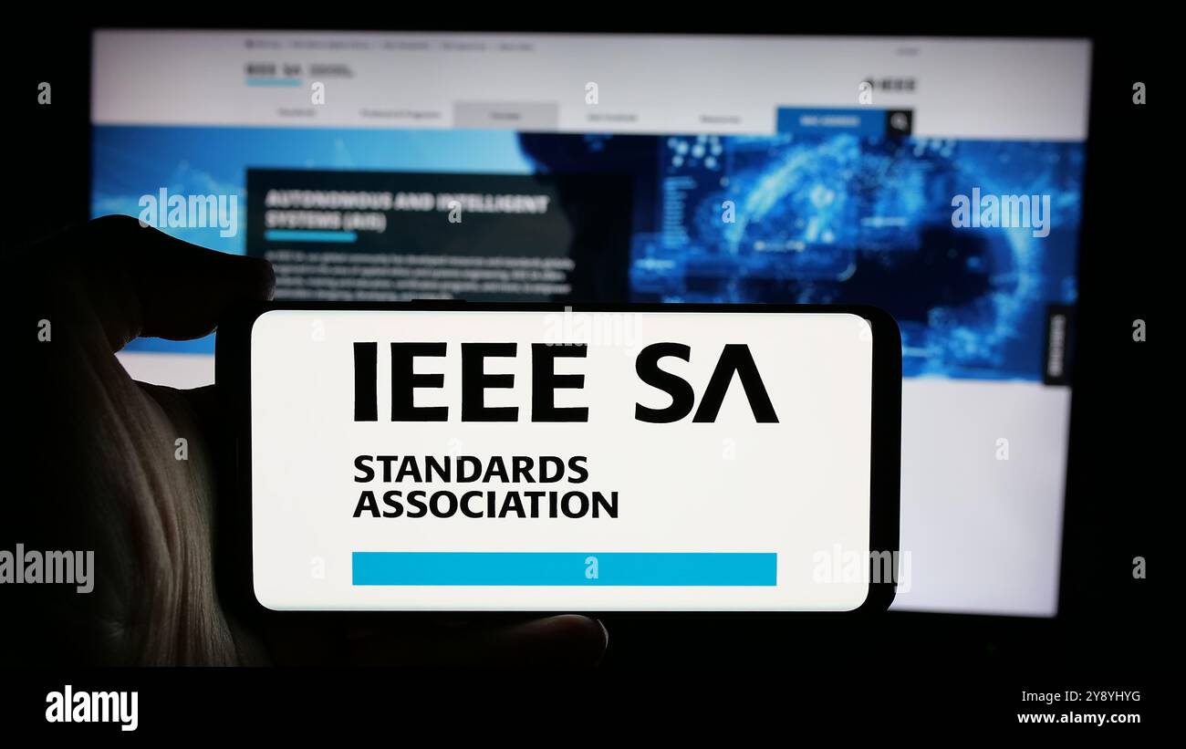Persona che possiede uno smartphone con il logo della IEEE Standards Association sullo schermo di fronte al sito Web. Mettere a fuoco il display del telefono. Foto Stock