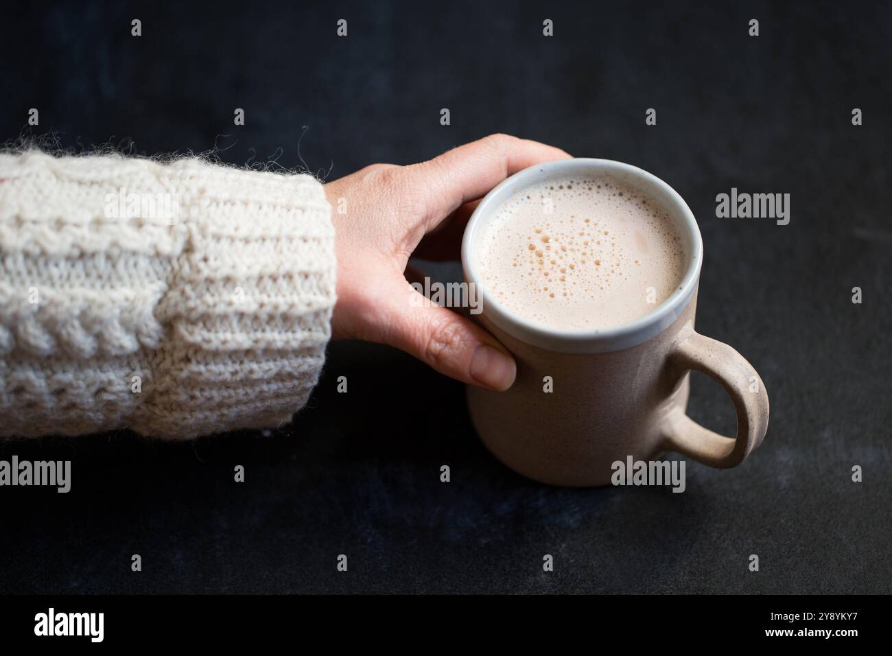 La mano di una donna che si avvicina per prendere una tazza di cioccolata calda. La donna indossa un ponticello a maglia. La tazza è in ceramica e la cioccolata calda è schiuma. Foto Stock