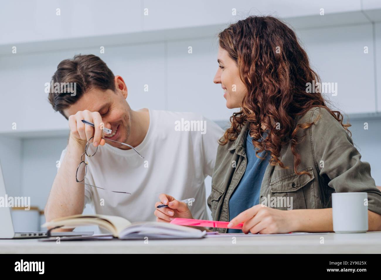 Coppia felice che lavora insieme a casa, ridendo e discutendo, lavoro di squadra informale e collaborazione. Uomo e donna sorridenti circondati da libri e notebook, godendosi momenti spensierati. Foto Stock