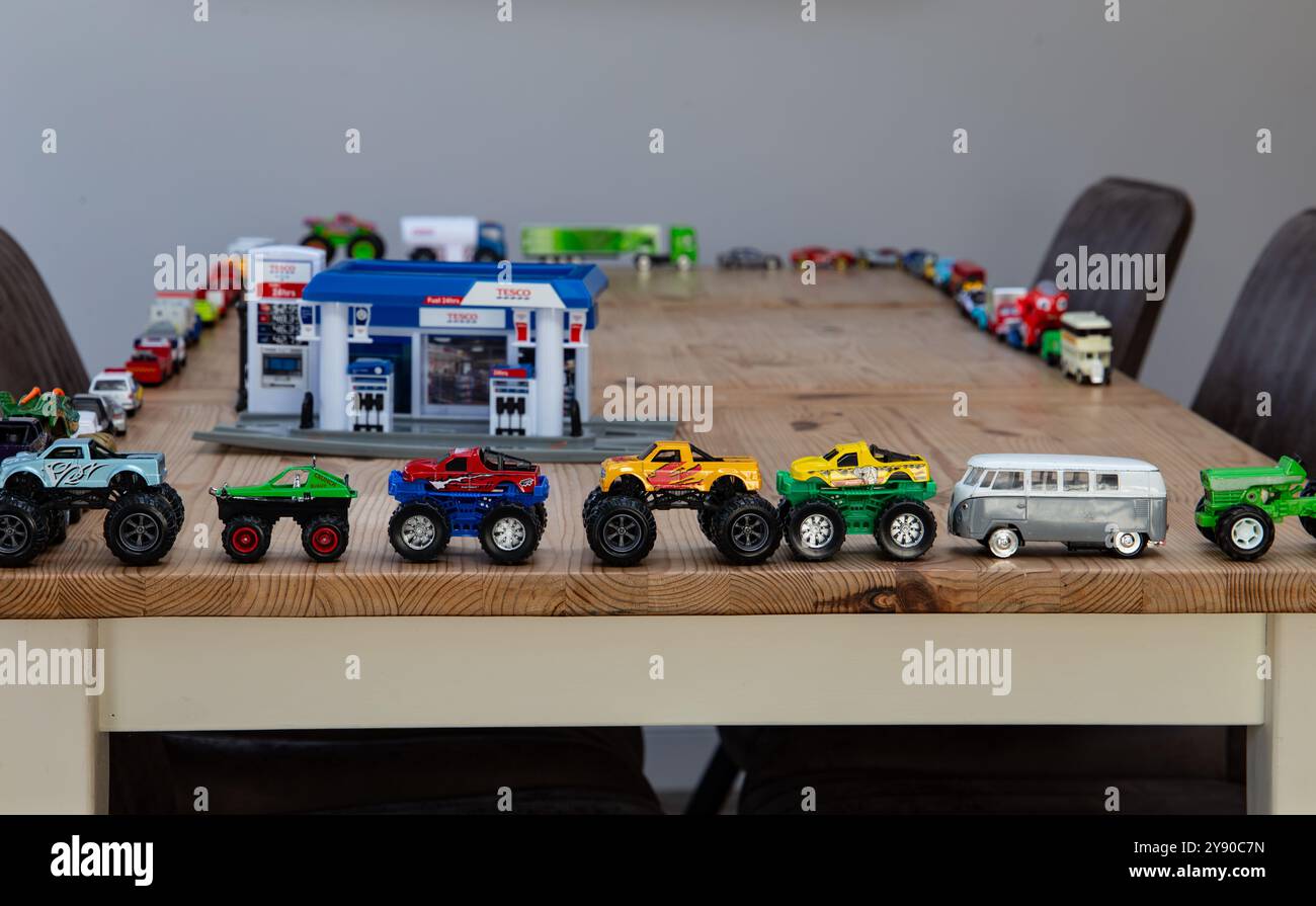 Una collezione di macchinine colorate allineate su un tavolo di legno con una stazione di servizio/benzina giocattolo e altre auto sullo sfondo Foto Stock